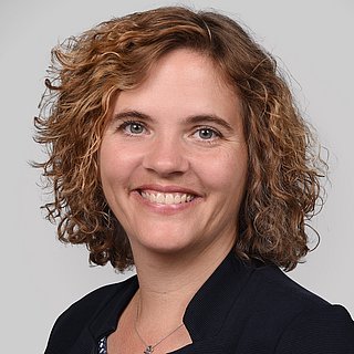 Eine weiße Frau mittleren Alters mit hellbraunen, lockigen, schulterlangen Haaren lächelt freundlich in die Kamera.