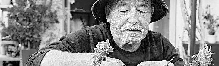 Schwarz-weiß Bild. Zu sehen ist ein alter Mann mit Sonnenhut. Seine Hände arbeiten in der in einem Blumenkasten